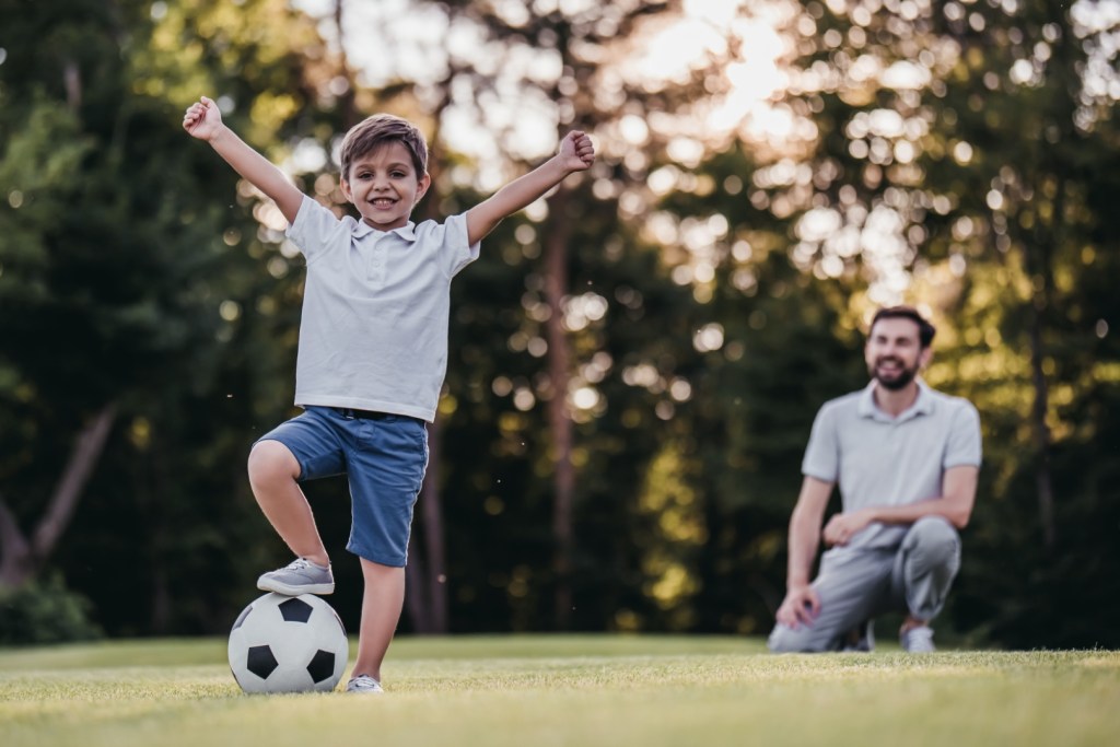 Chłopiec wznosi w górę ręce w geście triumfu podczas gry w piłkę nożna z tatą w parku